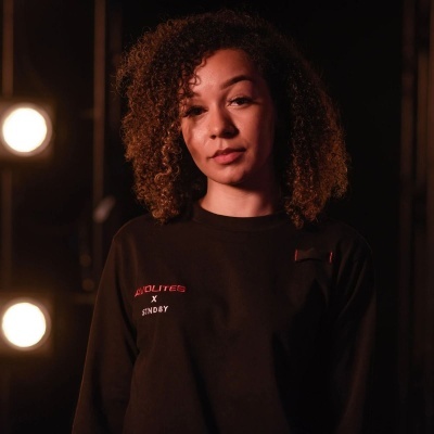 Mulher com camisola preta bordada com texto, em ambiente iluminado com luzes desfocadas ao fundo