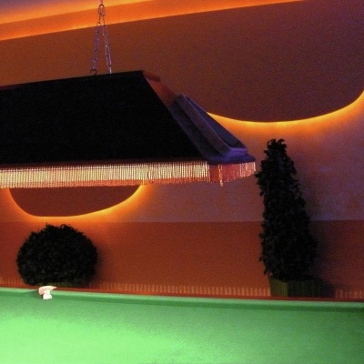 Mesa de bilhar verde sob iluminação com franja pendente, parede laranja com luzes LED em formato ondulado e plantas em vasos