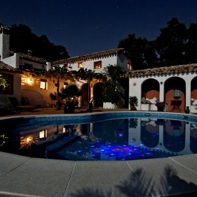 Piscina redonda iluminada à noite com casa branca e telhado de telhas castanhas ao fundo