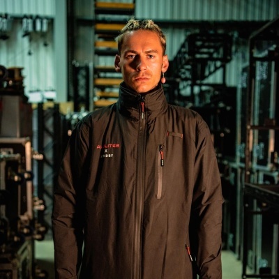 Casaco preto com fecho de correr e texto bordado, usado por homem em ambiente industrial