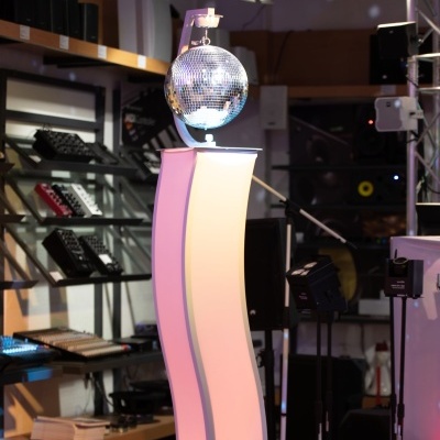 Esfera de discoteca espelhada num suporte branco curvo com iluminação em estúdio e equipamento musical ao fundo