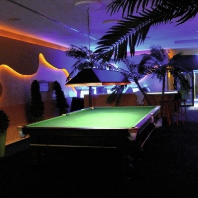 Mesa de bilhar com lona verde num ambiente iluminado com parede laranja e plantas