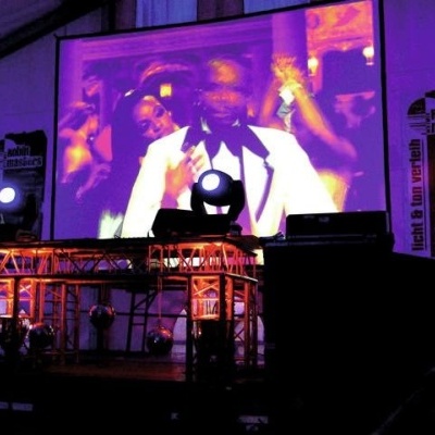 Projeção de pessoa em fato branco num palco com iluminação roxa e cartazes ao fundo