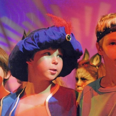 Quatro crianças em ambiente iluminado com luz roxa, uma delas usa chapéu azul com pena vermelha.