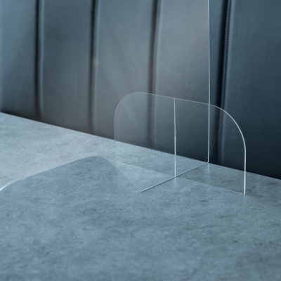 Separador de acrílico transparente sobre mesa cinza, com cadeiras pretas ao fundo