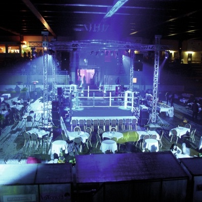 Ringue de boxe iluminado rodeado por mesas e cadeiras em evento interior