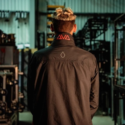 Casaco preto com gola vermelha e texto AVA, visto de costas em ambiente industrial.