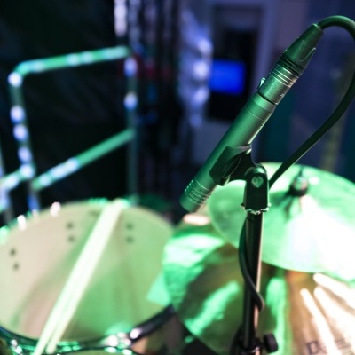 Microfone preto em suporte junto de bateria acústica com prato metálico e pele branca, luz verde e azul