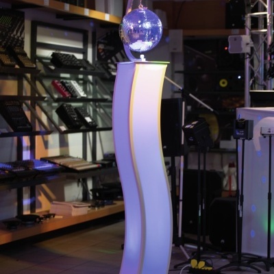 Coluna iluminada branca curvada com bola de espelhos tipo discoteca prateada no topo, num espaço interior com prateleiras e equipamentos