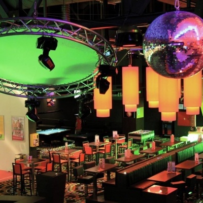 Interior de bar ou discoteca com mesas, cadeiras, iluminação verde e laranja, e globo espelhado suspenso