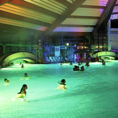 Piscina interior com iluminação verde, pessoas a nadar e teto com vigas altas