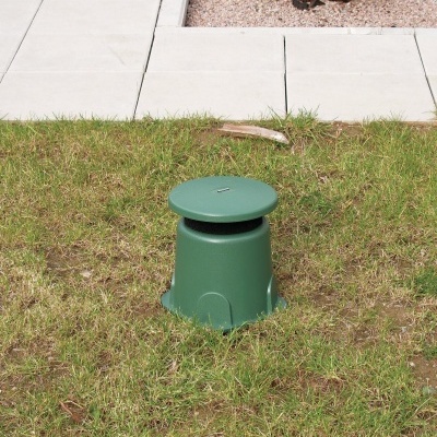 Elemento verde de plástico com tampa circular na relva junto a passeio de pedras e cascalho