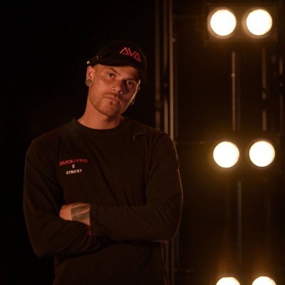 Homem com gorro e camisola preta com letras vermelhas e luzes ao fundo