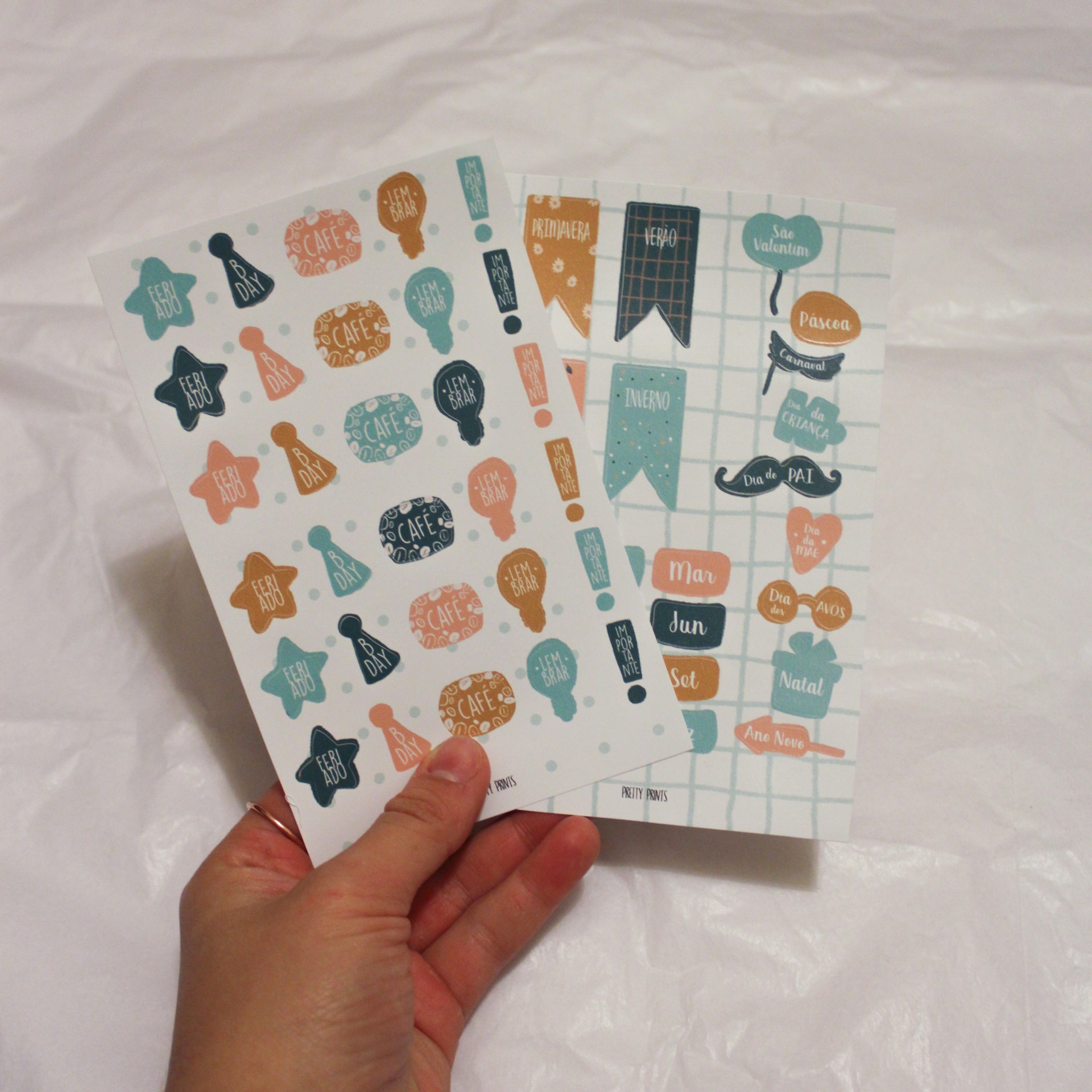 Dois conjuntos de autocolantes decorativos coloridos em papel, segurados por uma mão, sobre fundo branco