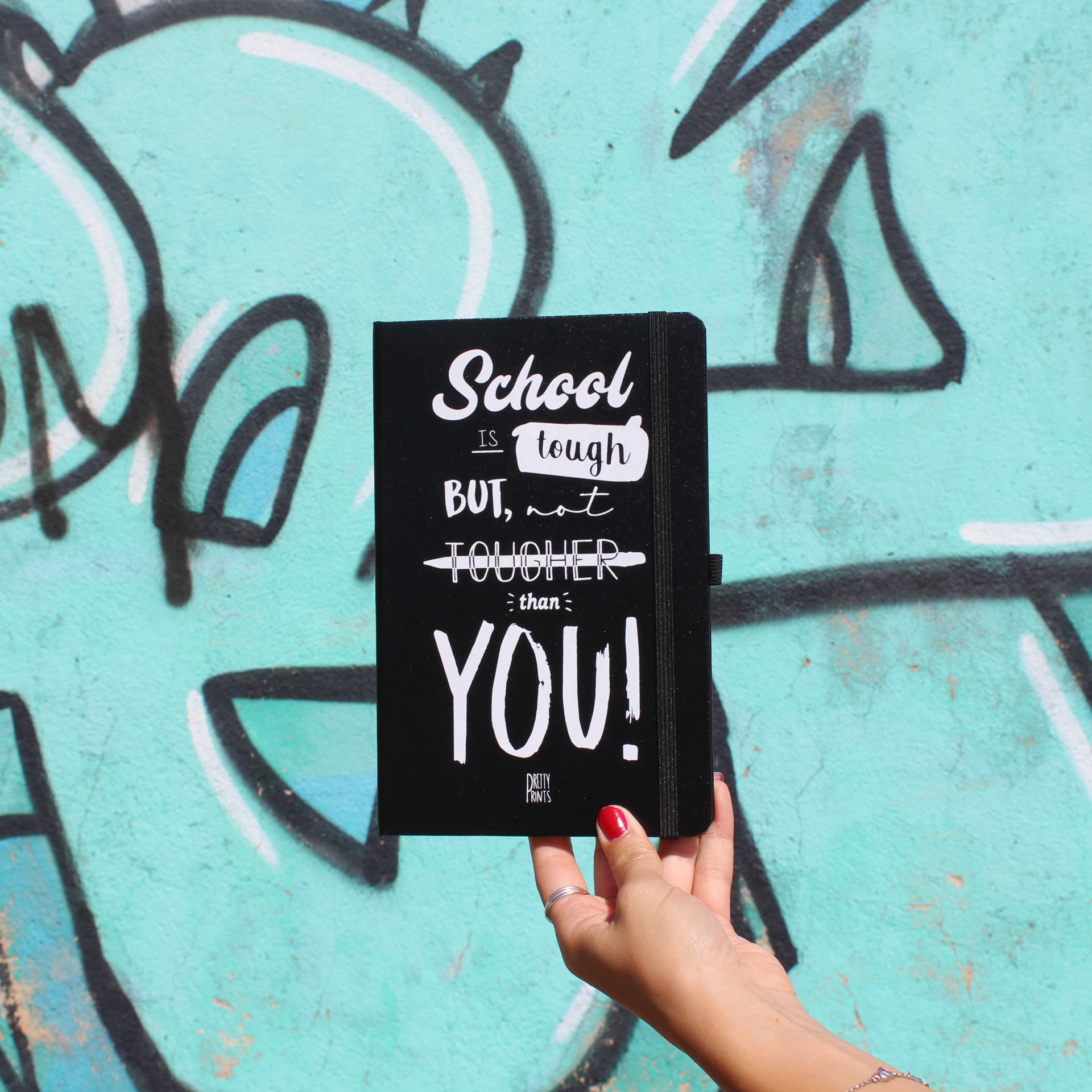Caderno preto com frase em branco segurado por mão com unhas vermelhas frente a parede com graffiti azul