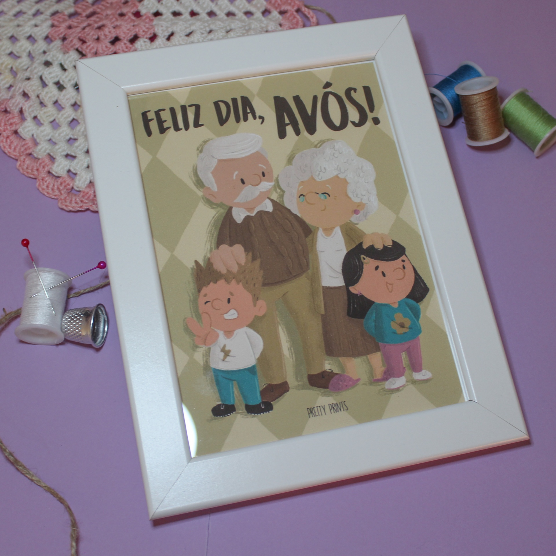 Moldura branca com ilustração de avós e netos e texto FELIZ DIA, AVÓS!, sobre superfície lilás com materiais de costura