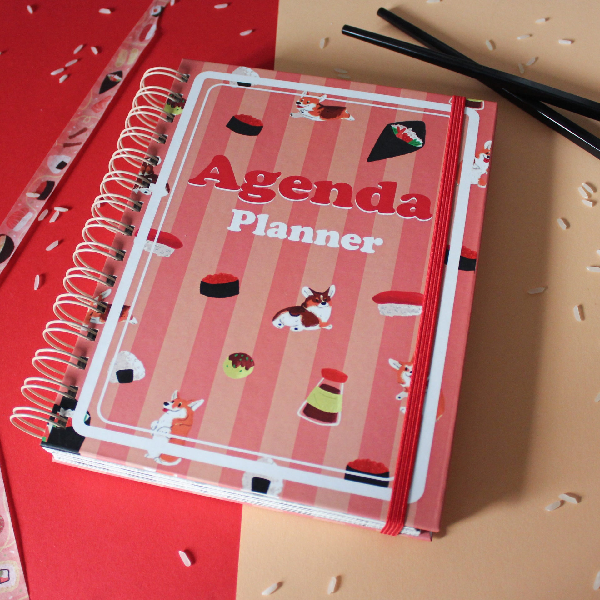 Agenda planner com capa ilustrada em tons de rosa, riscas e desenhos de sushi e cães, com espiral e elástico vermelho