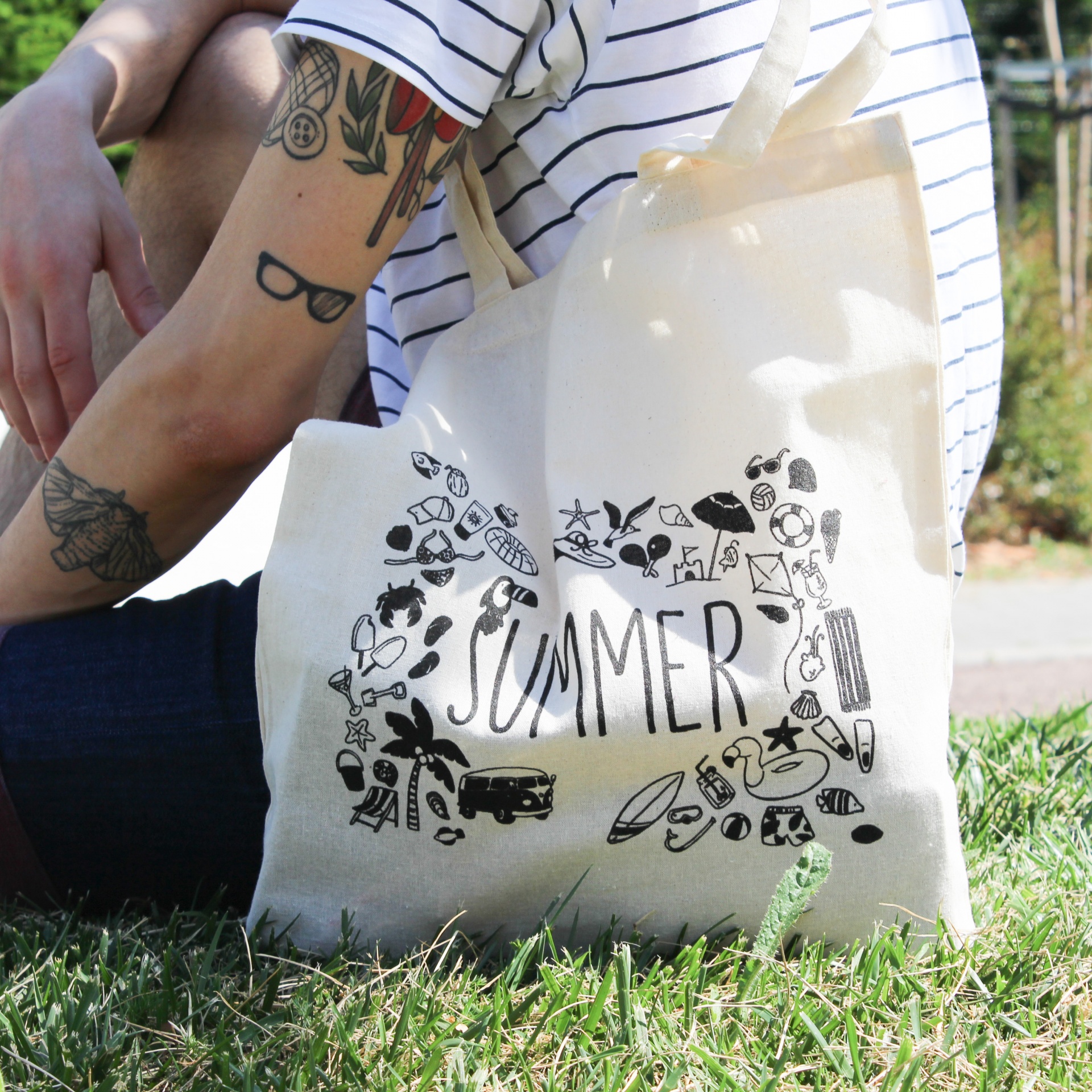 Saco de tecido com ilustração e palavra SUMMER, apoiado na relva junto a pessoa com t-shirt às riscas