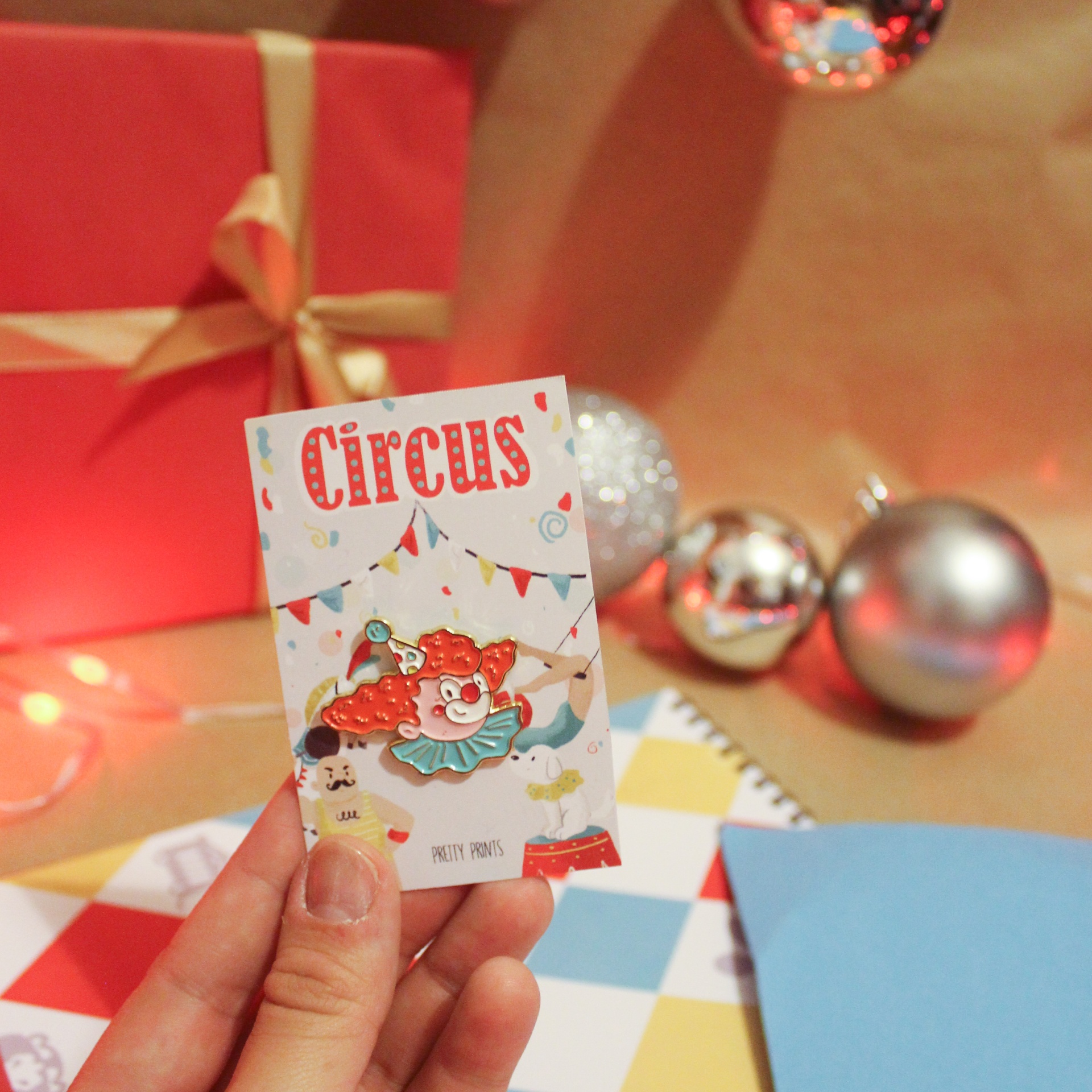 Cartão pequeno colorido com palhaço e texto Circus segurado à mão com unhas roxas sobre fundo festivo com bolas e caixa