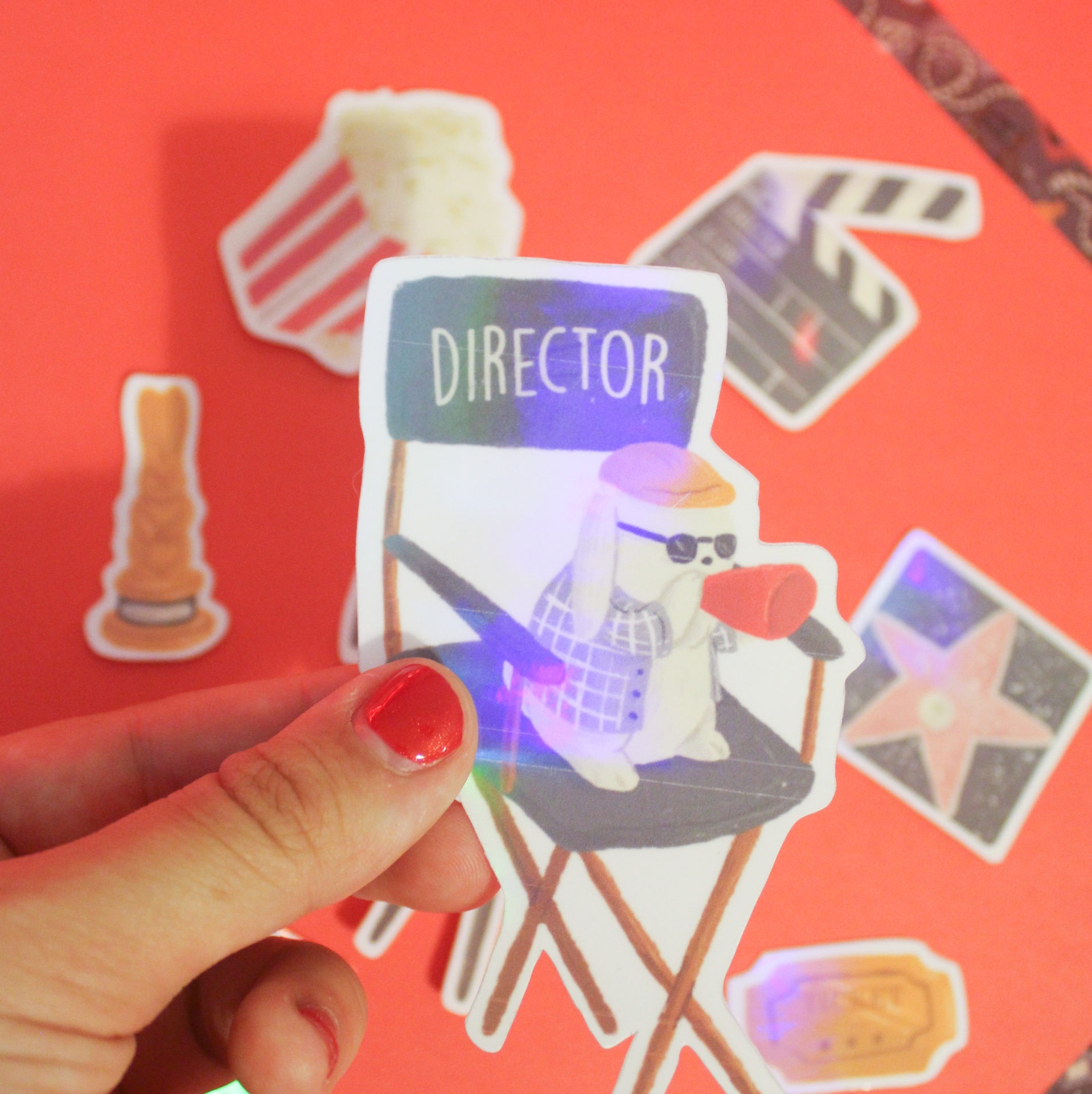 Autocolante holográfico de personagem sentado numa cadeira de director com a palavra DIRECTOR, rodeado por autocolantes de cinema sobre fundo vermelho.