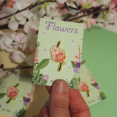 Broche em forma de flor dourado e rosa em cartão verde com texto Flowers e ilustrações florais, segurado por mão