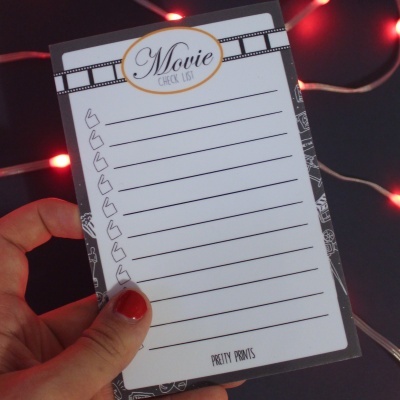 Bloco de lista para filmes com design em preto e branco sendo segurado por mão com unhas vermelhas