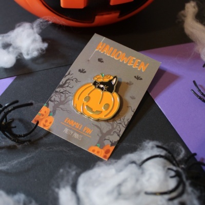 Pin esmaltado de Halloween com abóbora e gatinho sobre cartão cinza, com aranhas e teia falsa