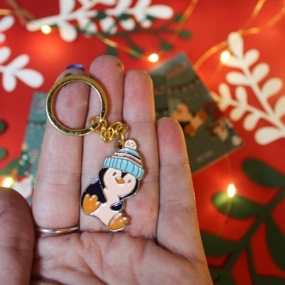 Porta-chaves com pinguim de gorro azul claro e pompom, em mão sobre fundo natalício vermelho com luzes e decoração branca e verde.