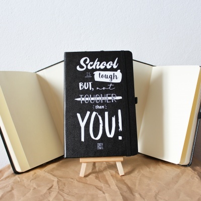 Caderno preto com texto branco em suporte de madeira sobre papel castanho com dois cadernos abertos ao fundo