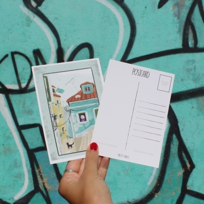 Duas postais seguradas por uma mão feminina com unhas vermelhas, com fundo grafitado turquesa