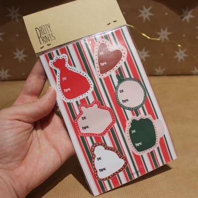 Etiqueta de presente com desenhos natalícios vermelhos, verdes e brancos, embalagem com aba castanha e logo 'PRETTY PRINTS'.