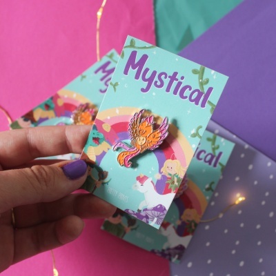 Cartão colorido verde-água com ilustração e texto 'Mystical' a segurar um pin esmaltado de ave mitológica.