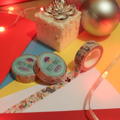 Rolos de fita adesiva colorida com padrões variados, caixa decorativa branca com enfeite prateado e bola dourada em fundo colorido com luzes LED.