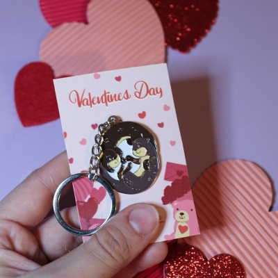 Porta-chaves metálico com casal de lontras e cartão 'Valentines Day' em fundo com corações