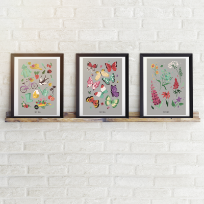 Três quadros coloridos com desenhos de bicicletas, borboletas e flores emoldurados numa prateleira frente a parede branca