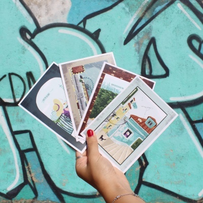 Mão a segurar cinco postais ilustrados em fundo de graffiti azul