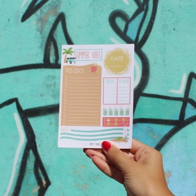 Bloco de notas 'Summer Vibes' com listas e ilustrações de verão, segurado com mão, fundo de parede azul com grafite