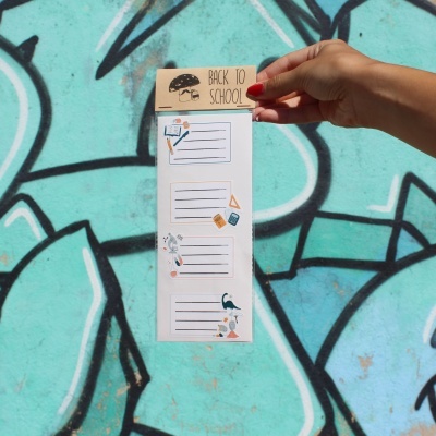 Bloco de anotações 'BACK TO SCHOOL' com desenhos escolares, segurado por mão, fundo grafite azul