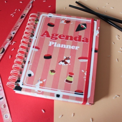 Agenda Planner rosa com desenhos de sushi, elástico vermelho e paus de comida pretos sobre fundo vermelho e bege