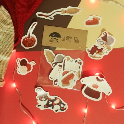 Autocolantes de papel temáticos de outono com ilustrações variadas sobre mesa colorida com manta e luzes.