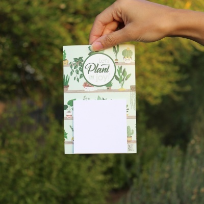 Bloco de notas com plantas e texto 'Let's Plant Joy' em fundo ao ar livre