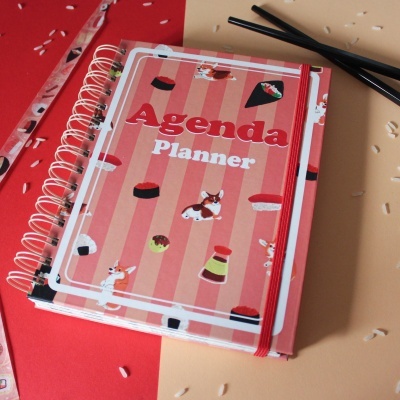 Agenda planner com capa ilustrada em tons de rosa, riscas e desenhos de sushi e cães, com espiral e elástico vermelho