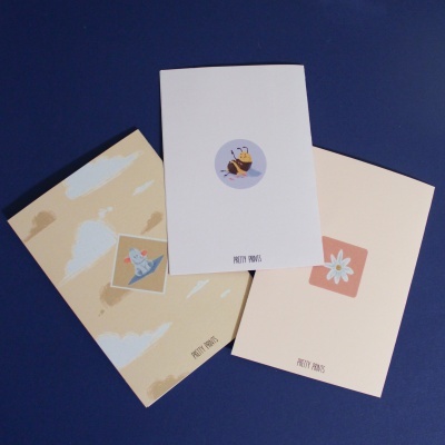 Três cadernos com capas em tons neutros e ilustrações simples, em fundo azul escuro.