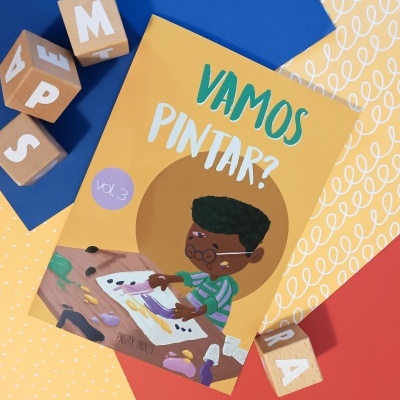 Livro infantil 'VAMOS PINTAR?' com criança a pintar e cubos de madeira coloridos ao redor