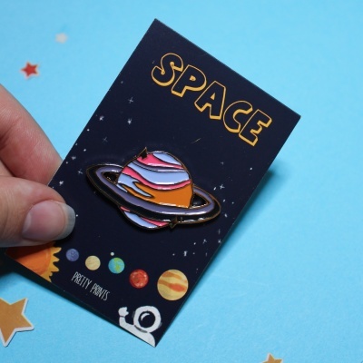 Pin esmaltado de planeta com anéis em cartão preto com texto SPACE e ilustrações espaciais