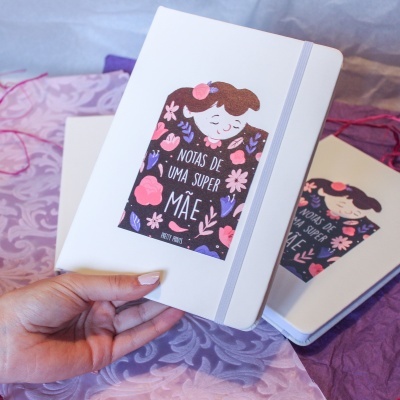 Caderno branco com ilustração e texto para mães sobre tecido lilás e rosa.