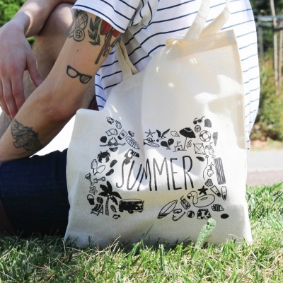 Saco de tecido com ilustração e palavra SUMMER, apoiado na relva junto a pessoa com t-shirt às riscas