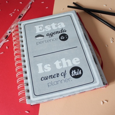 Agenda cinza espiral com texto em português e inglês sobre superfície vermelha e bege com palitos pretos e arroz.