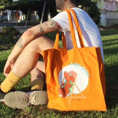 Saco laranja com desenho de cachorro quente e texto HOT DOG carregado por pessoa sentada na relva