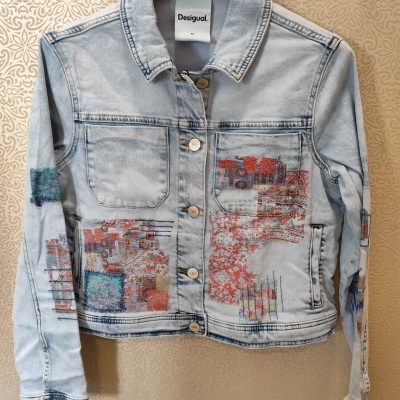 Casaco de ganga azul clara Desigual com estampados coloridos e botões metálicos pendurado num cabide preto